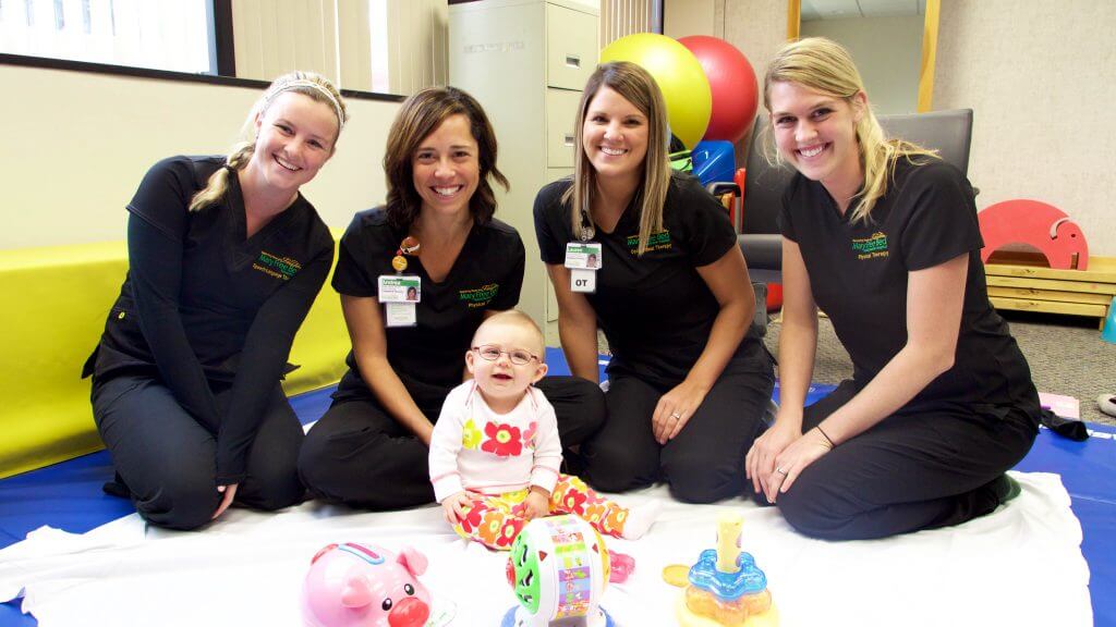 Baby wearing glasses sitting on blanket surrounded by her therapy team, four women wearing black uniforms