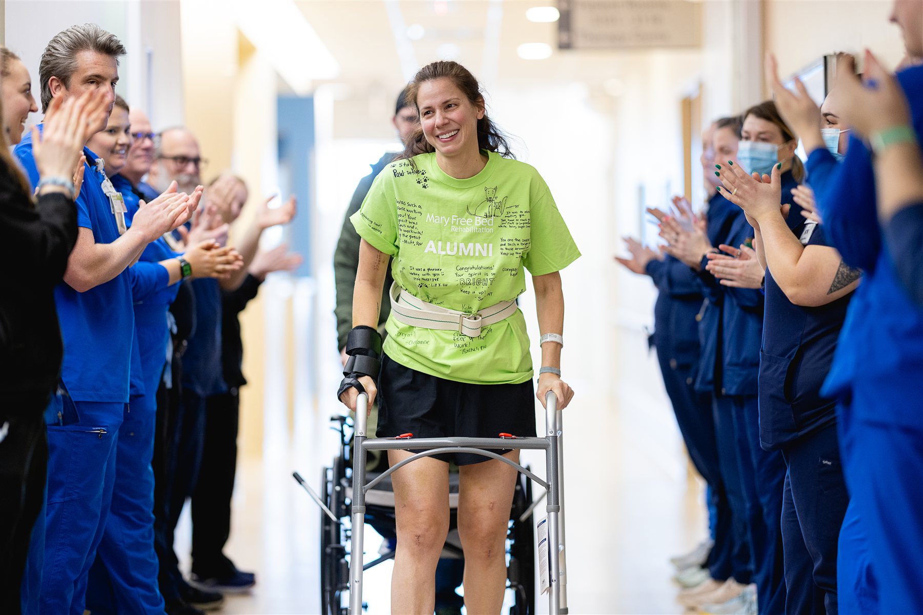 A patient in an alumni T-shirt walks with a walker down a hallway as Mary Free Bed team members applaud her graduation