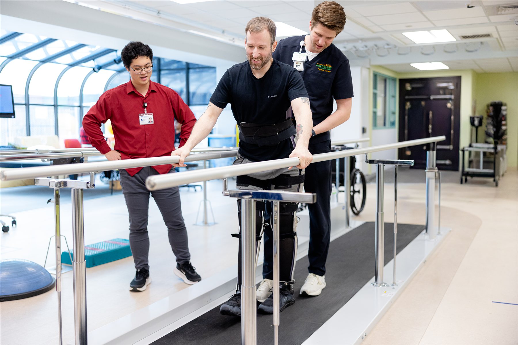 A patient walks between parallel bars in a robotic exoskeleton with two Mary Free Bed therapists at his side