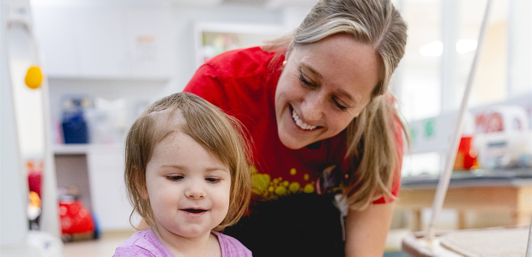 A young patient smiles on the therapy gym floor while her therapist leans in beside her