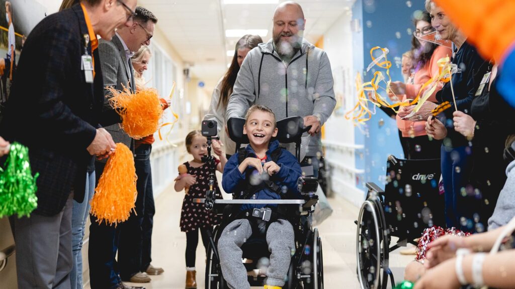 Staff and family cheer a young patient in a wheelchair down a hospital hallway