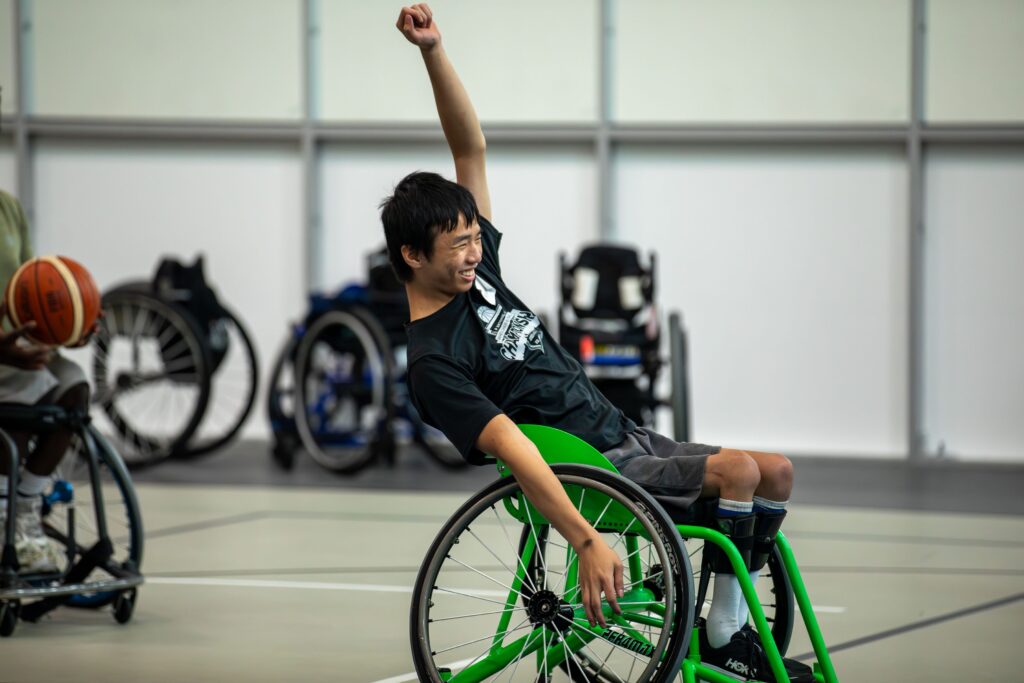A young athlete in a sport wheelchair raises his arm in celebration on the court