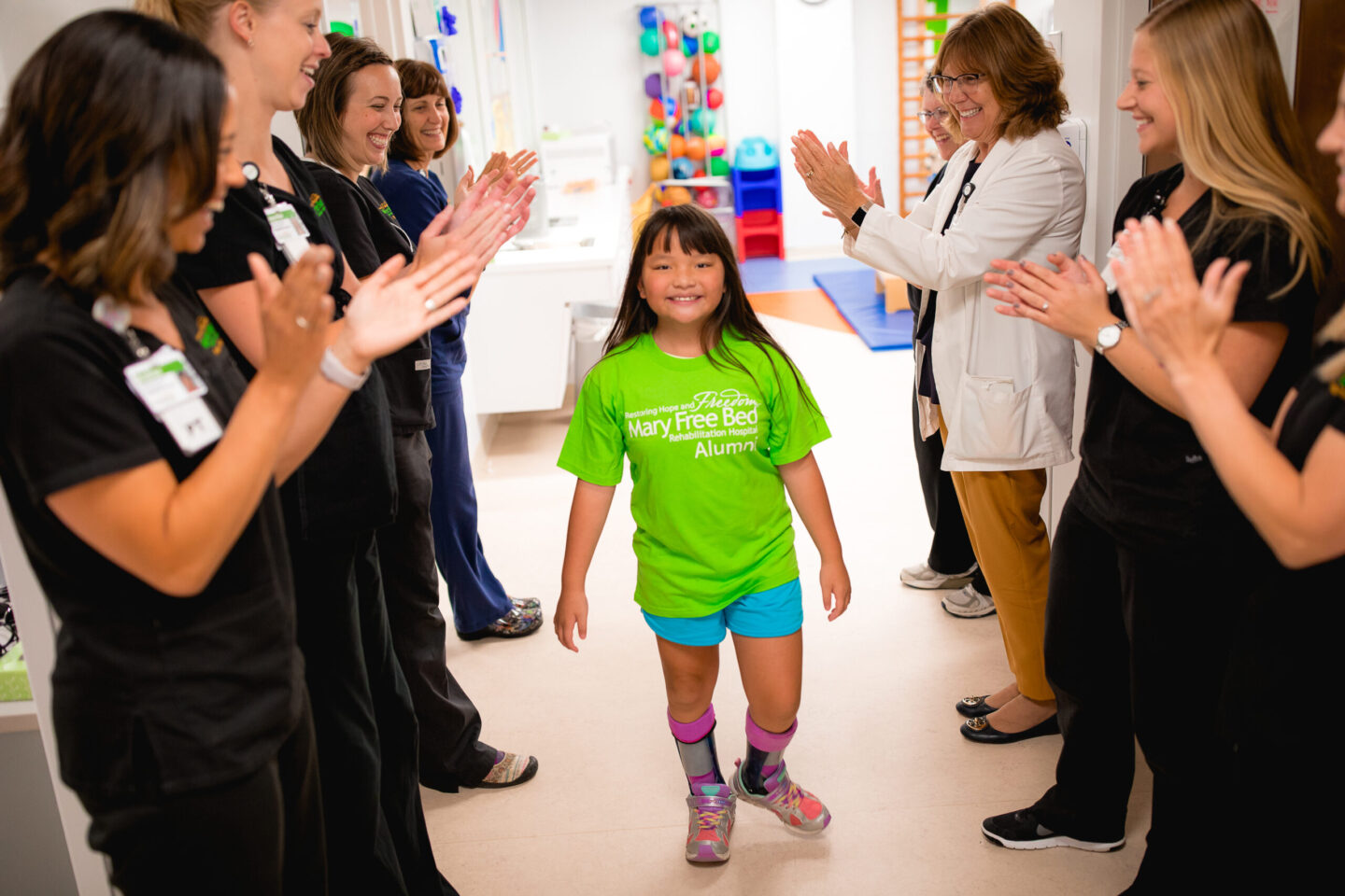 A young patient with leg braces walks confidently through a hallway as staff applaud her progress.