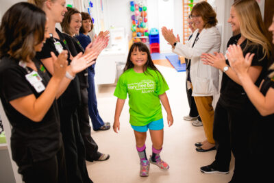 A young patient with leg braces walks confidently through a hallway as staff applaud her progress.