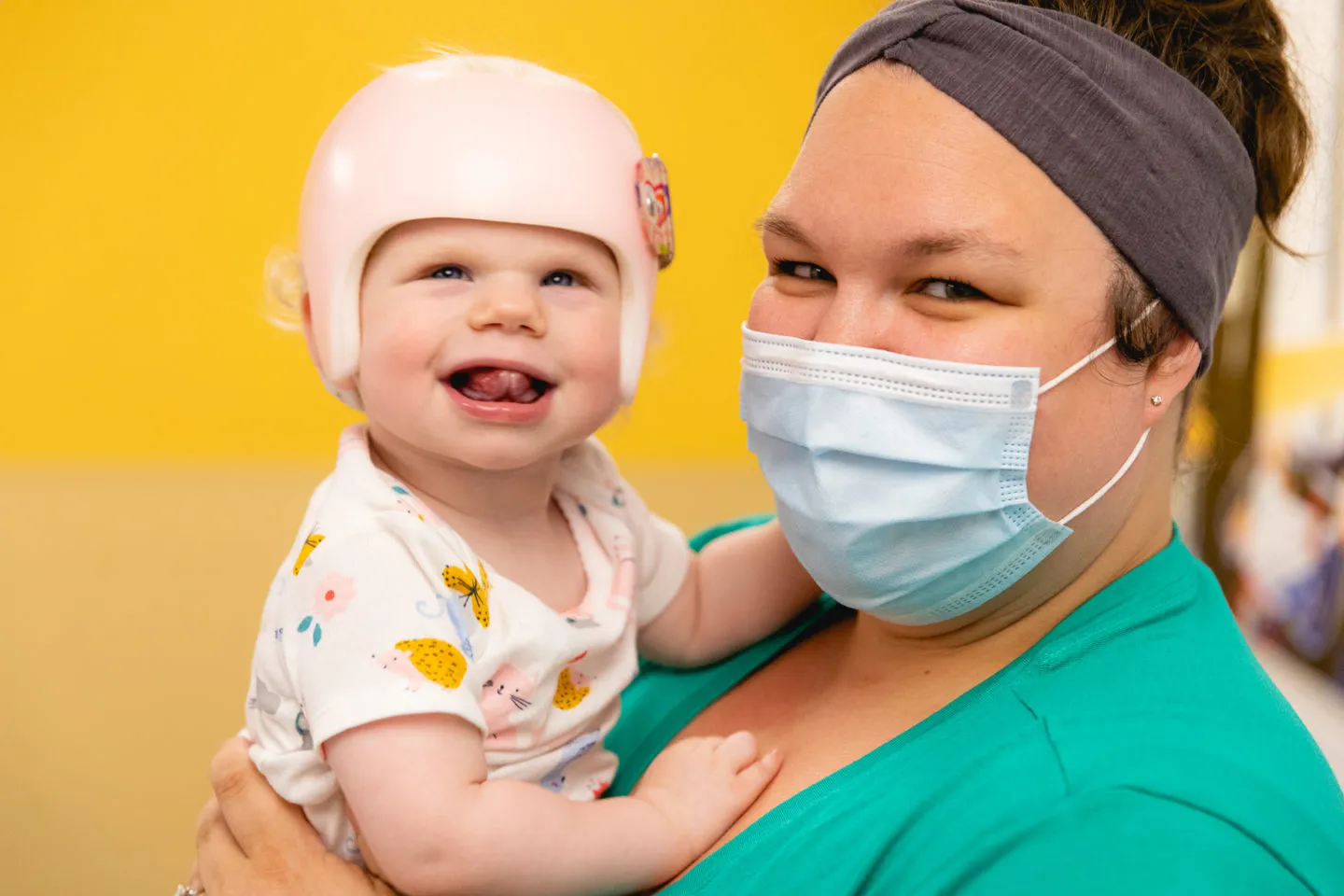 Smiling baby wearing a cranial helmet held by a masked pediatric healthcare provider in green scrubs