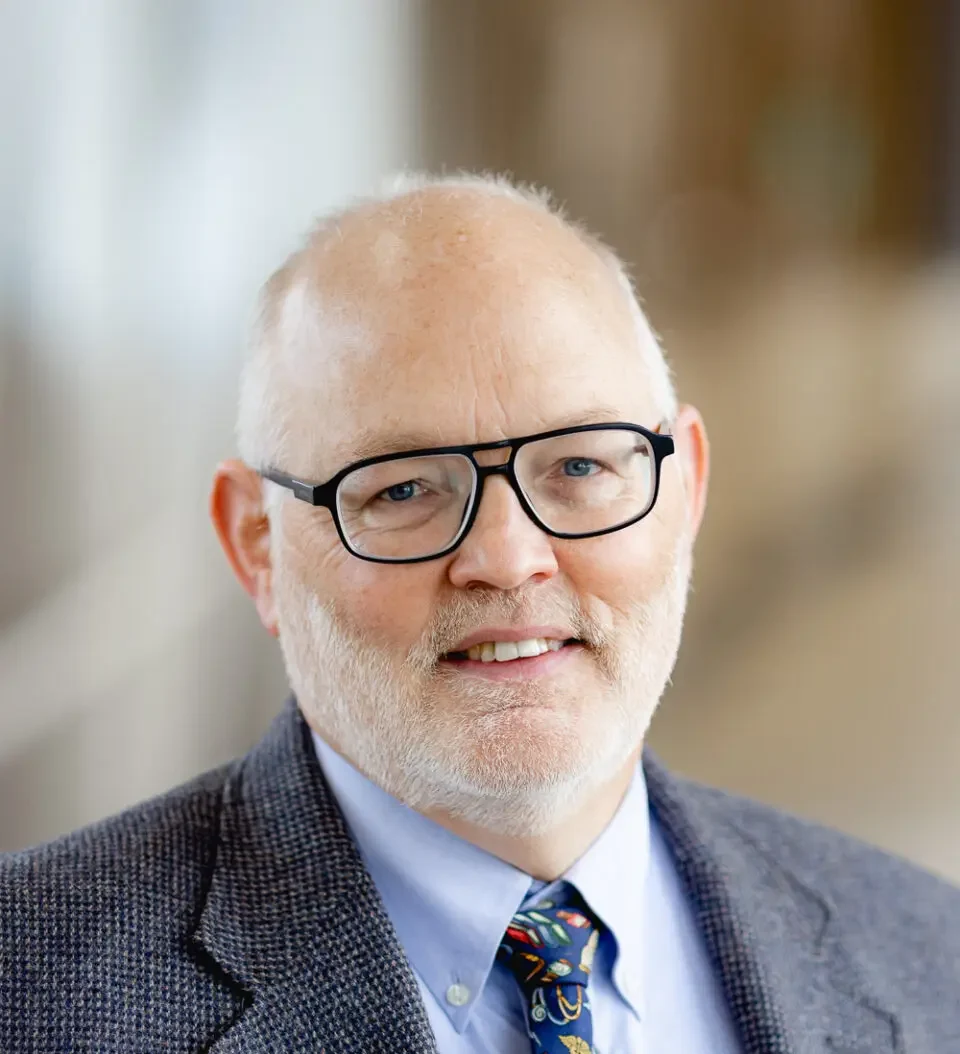Male pediatric physician smiling, wearing glasses, gray blazer, blue shirt, and medical-themed tie