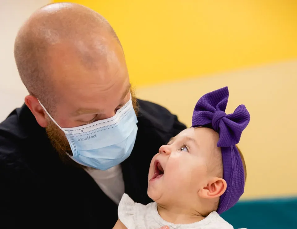 A masked pediatric therapist supports a smiling baby girl with a purple bow headband during a rehabilitation session at Mary Free Bed