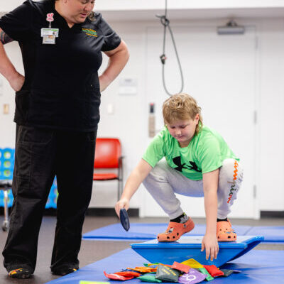 A pediatric therapist supervises a young child picking up colorful objects from a balance board during a rehab therapy session