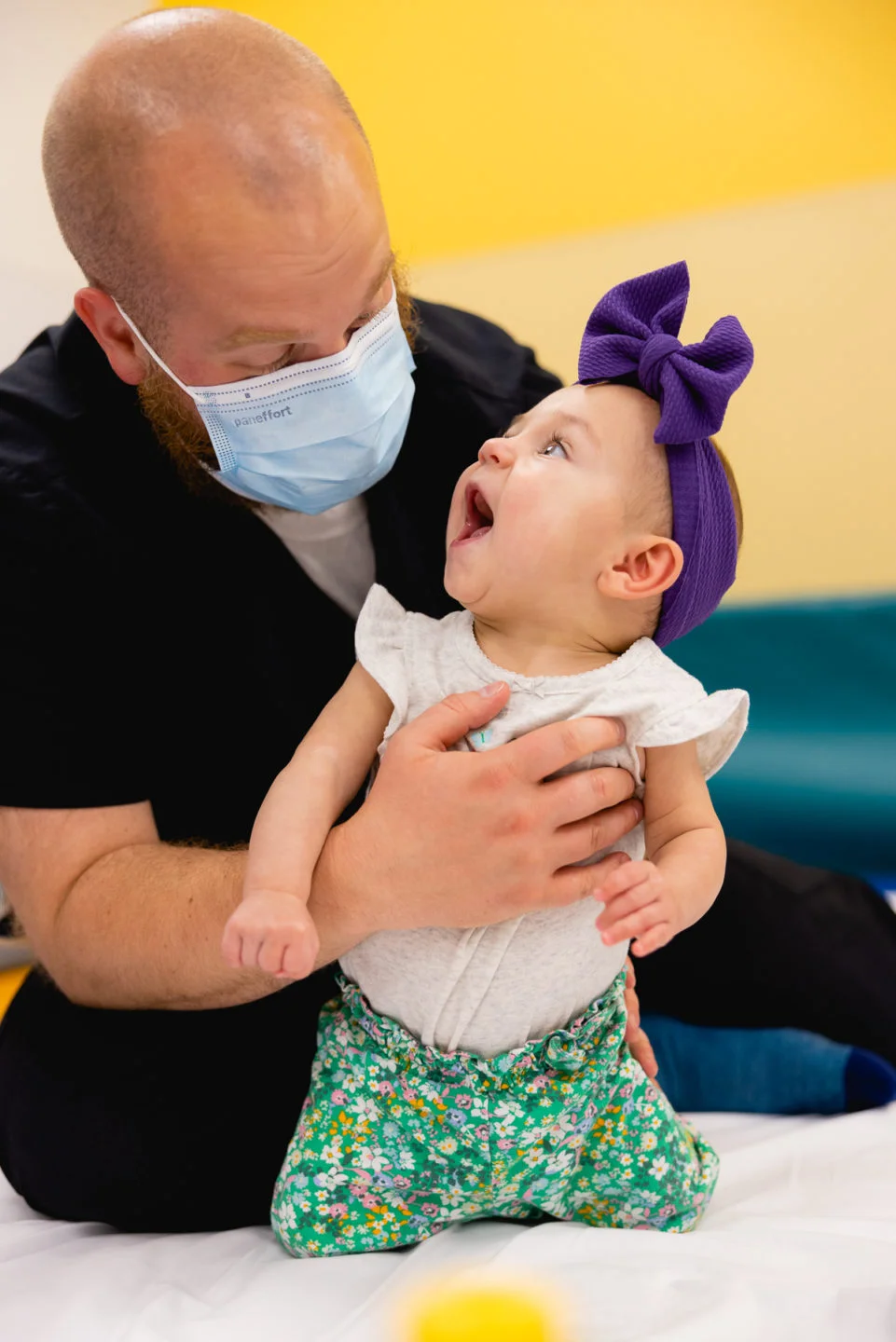 A masked pediatric therapist gently supports a smiling baby girl in a purple bow headband during a rehabilitation session