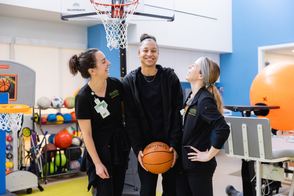 A teen patient holds a basketball with two smiling Mary Free Bed therapists in a pediatric rehabilitation gym with a basketball hoop