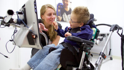 Young boy in adaptive stander touches therapist's face during pediatric rehabilitation session at Mary Free Bed