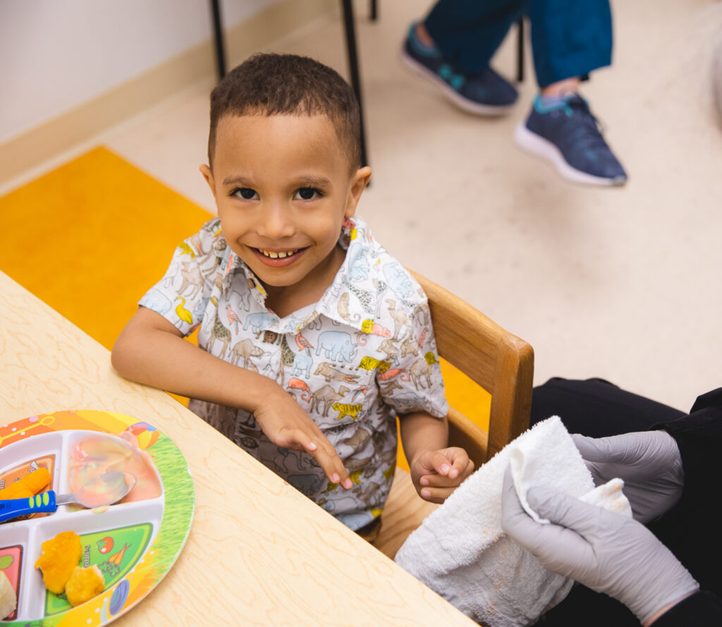 Smiling young boy in animal-print shirt sits at a table while a masked pediatric therapist assists with his bandaged hand during a meal session