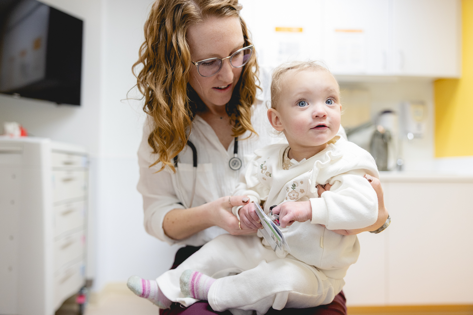 A pediatric clinician with a stethoscope gently holds and engages with a young baby during a clinic visit