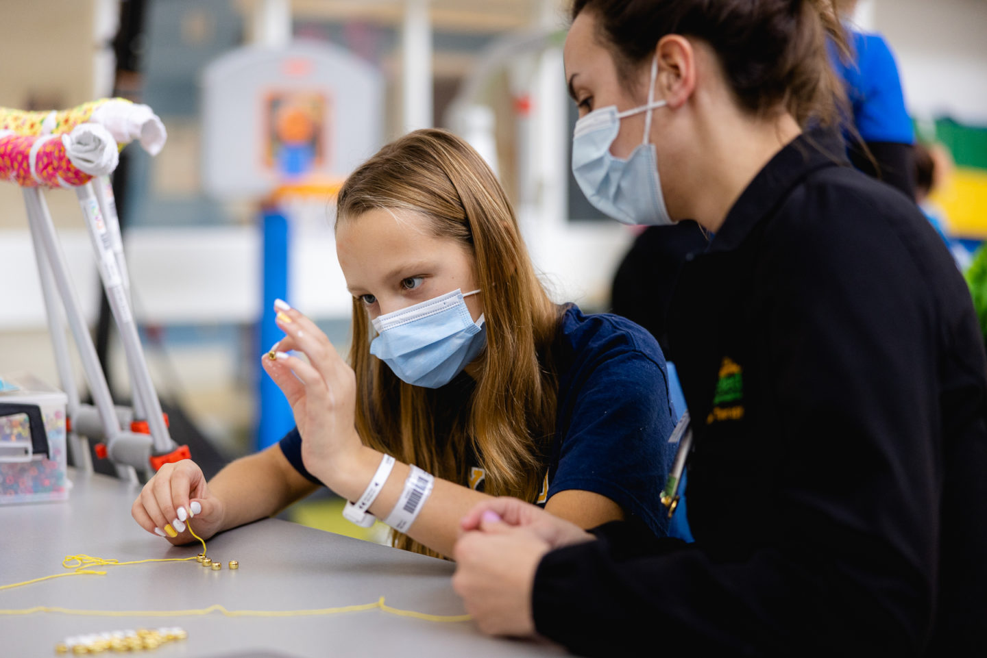 A masked pediatric therapist guides a young girl with crutches through a fine motor bead-stringing activity in a rehab gym