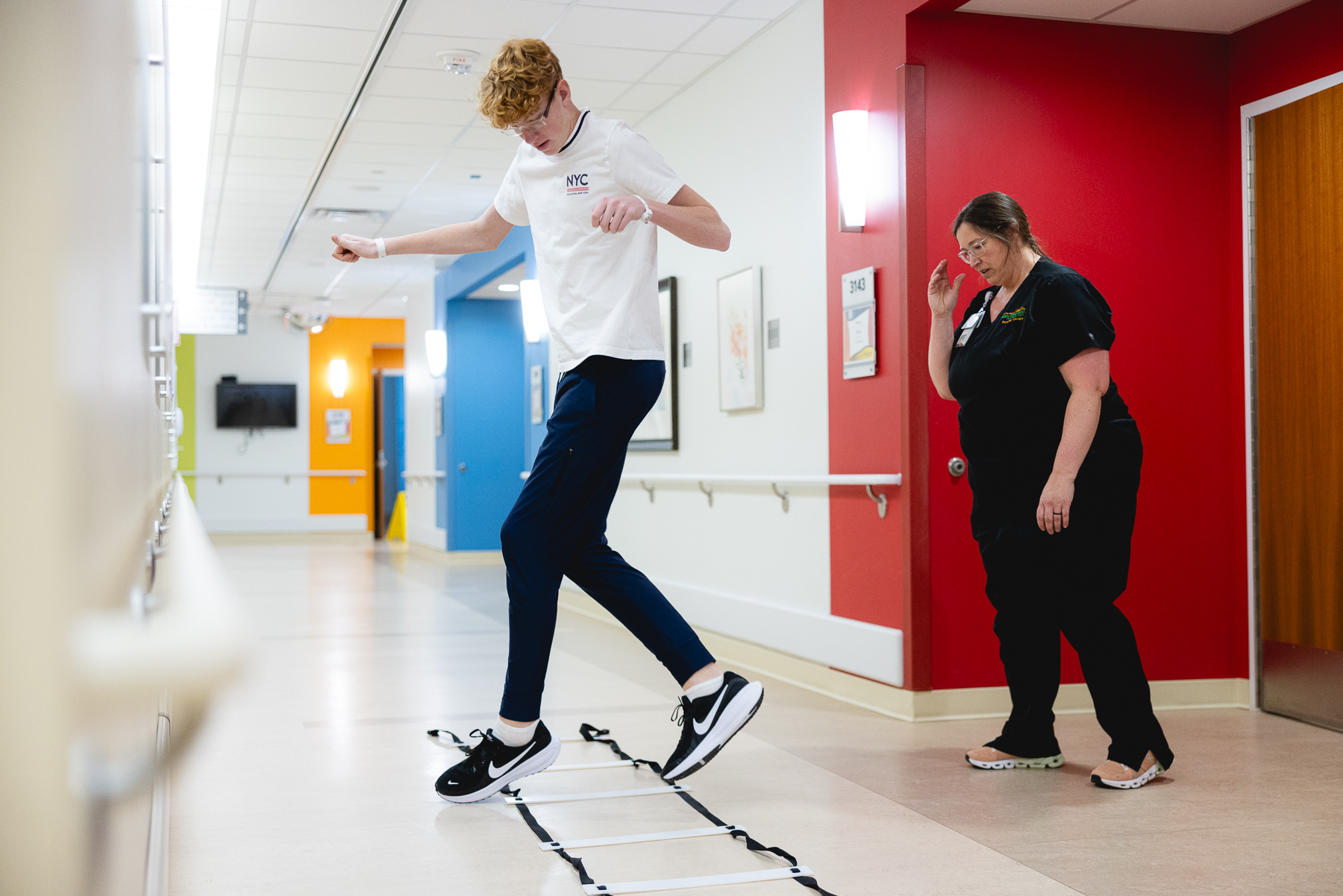 Pediatric physical therapist supervising a young patient performing jumping and coordination exercises on a ladder drill in a bright rehabilitation clinic, demonstrating motor skill development and athletic training