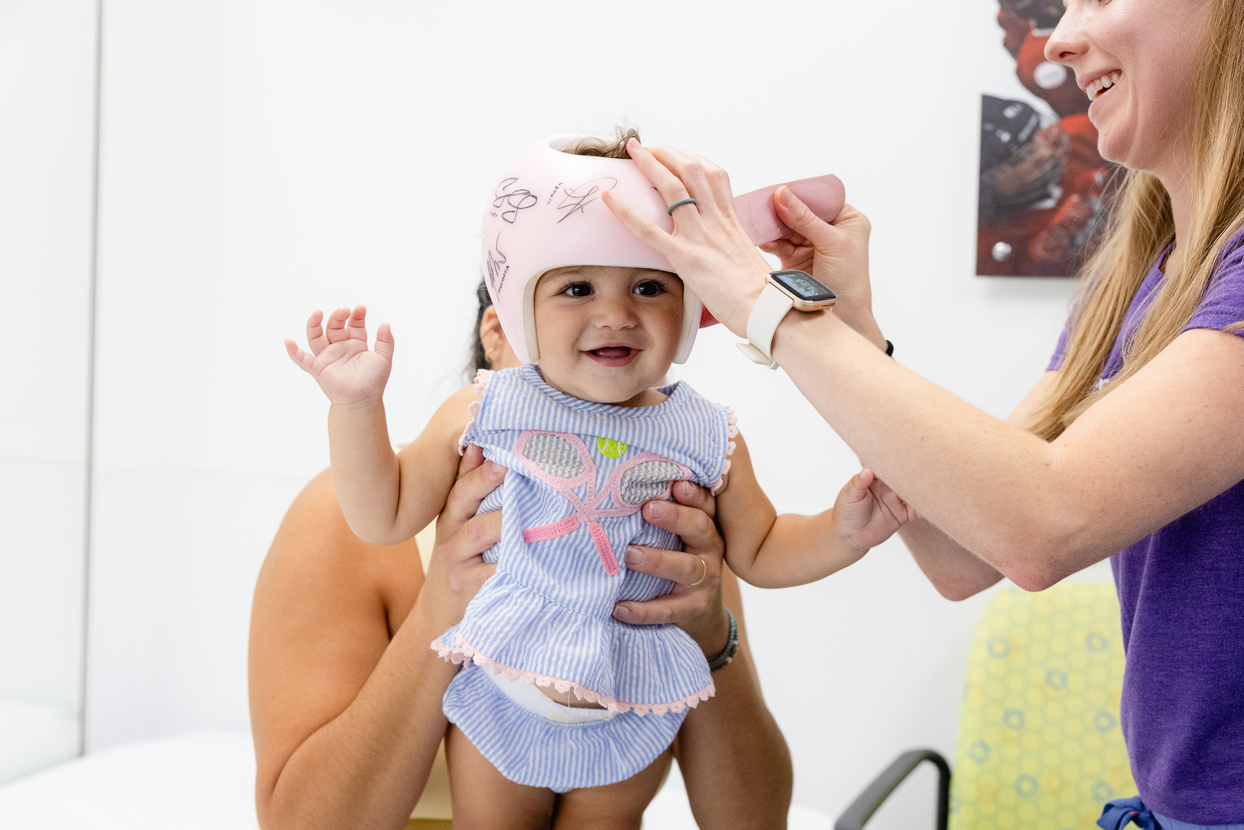 Smiling baby girl wearing a pink cranial helmet and blue seersucker romper, reaching her hands forward cheerfully
