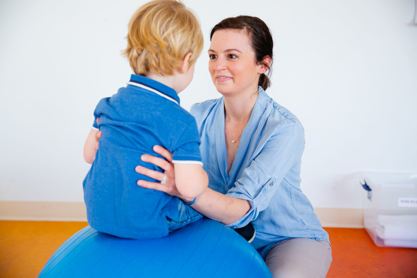 A pediatric therapist supports a young child sitting on a blue therapy ball during a rehabilitation session