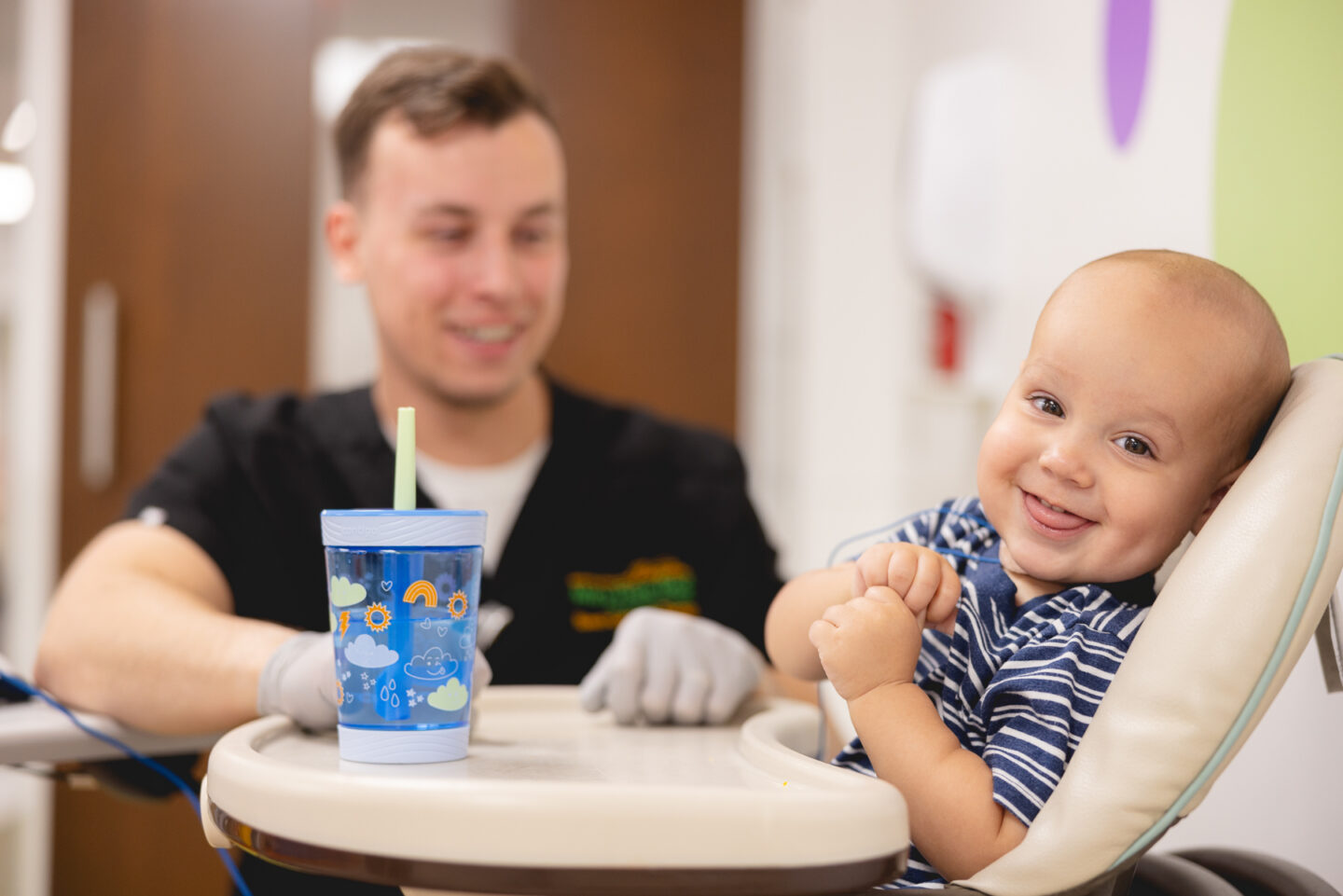 Smiling baby in a high chair during pediatric rehabilitation therapy with a Mary Free Bed therapist holding a sippy cup