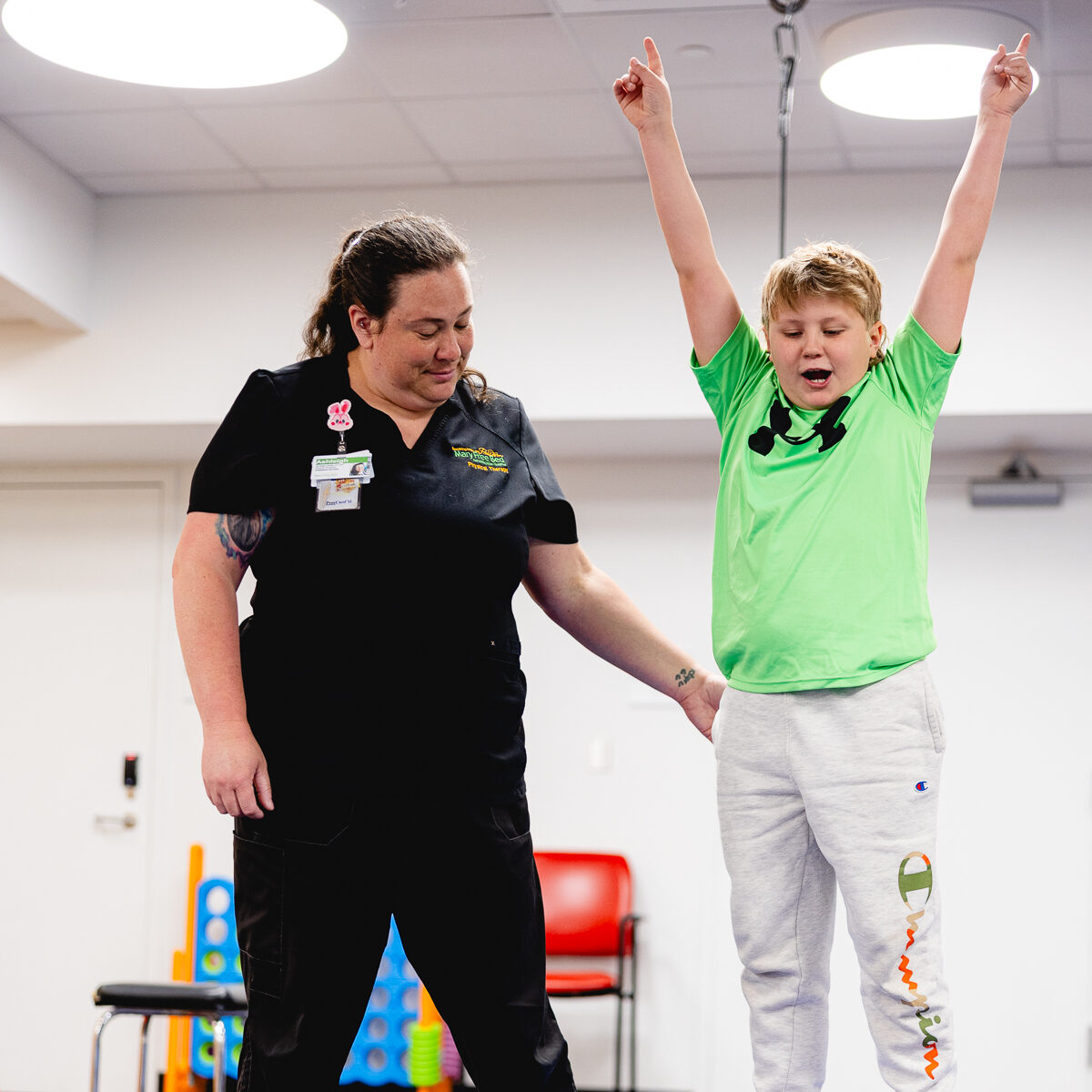 A Mary Free Bed pediatric therapist steadies a young boy as he celebrates with arms raised while balancing on a therapy board