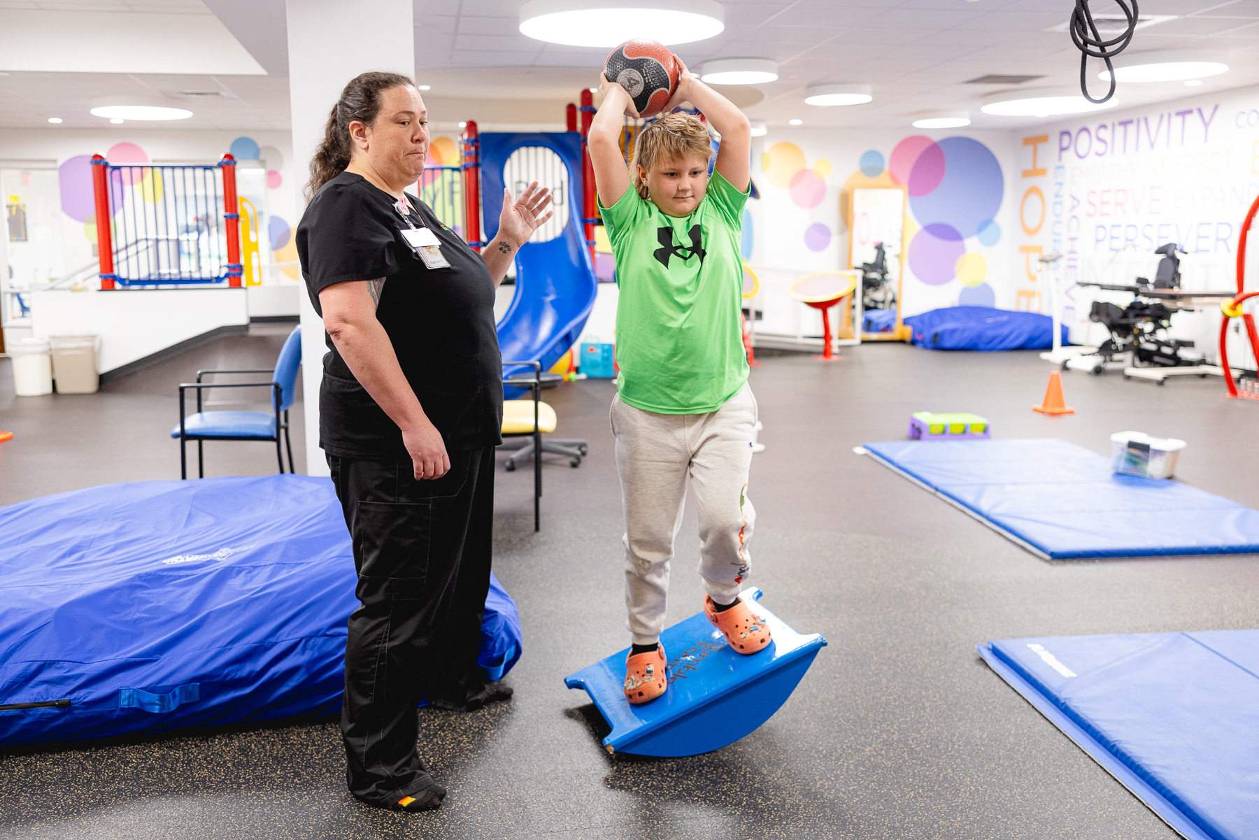 A pediatric therapist guides a child balancing on a rocker board while holding a medicine ball in a colorful rehab gym