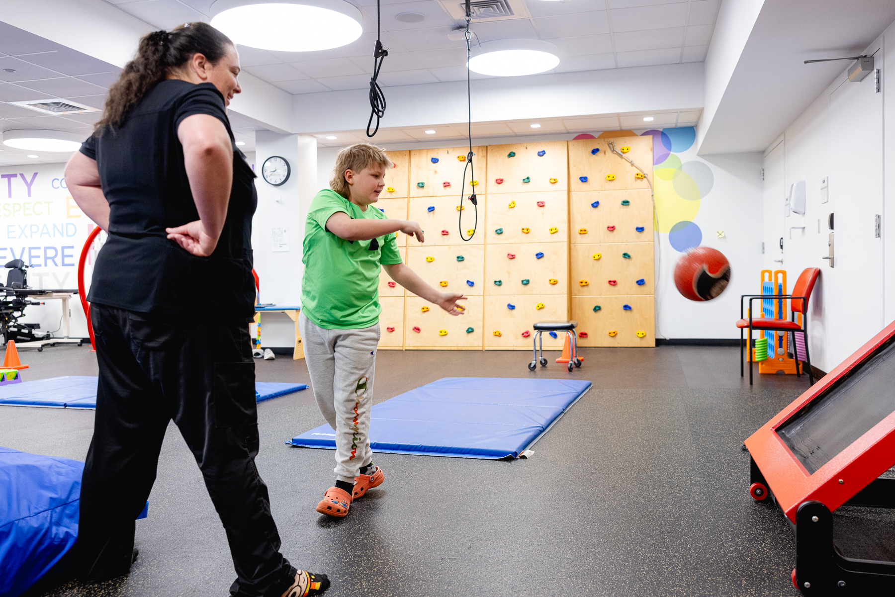A pediatric therapist guides a young boy through balance exercises in a colorful rehab gym with a climbing wall and therapy mats