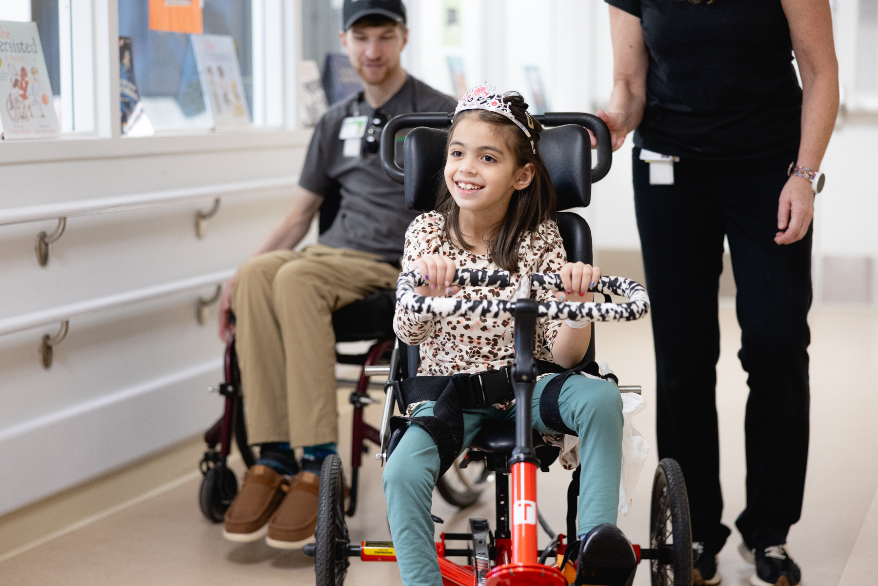 Smiling young girl wearing a tiara rides an adaptive tricycle during pediatric rehabilitation, supported by two Mary Free Bed therapists