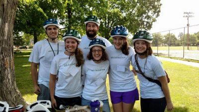 Group of six therapists wearing white t-shirts and bike helmets standing next to a tree with arms around each other