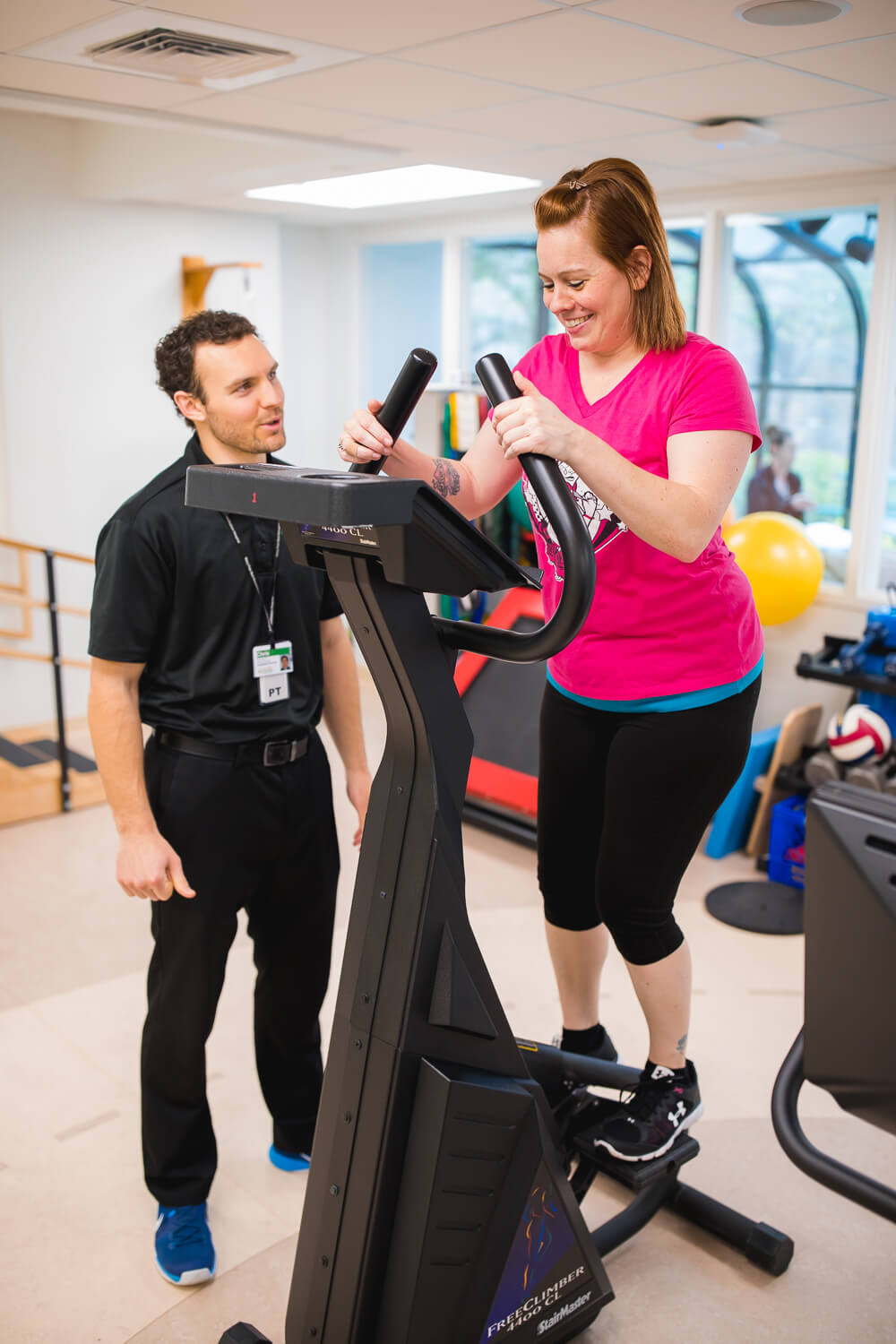A therapist is wearing a black uniform and talking to a female patient who is using a step machine, and wearing a bright pink shirt. They are in a gym setting.