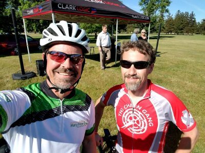 Two men in bicycle gear, one on the left is wearing a white bicycle helmet, and both men are wearing sunglasses and smiling outdoors. 