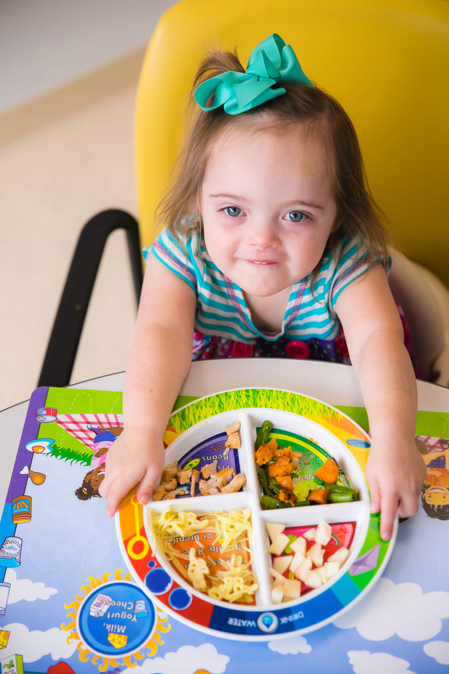 Pediatric Feeding Program patient - young girl with blue bow in her hair smiling and holding a colorful plate to the camera