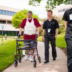- A male patient wearing a red t-shirt and turtle brace walks with a walker. A young male physical therapist wearing black uniform walks beside him on his right.