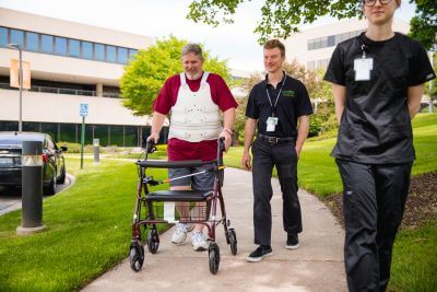- A male patient wearing a red t-shirt and turtle brace walks with a walker. A young male physical therapist wearing black uniform walks beside him on his right.