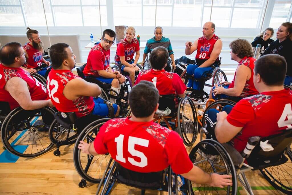 Mary Free Bed Sports Rehabilitation Program - A team of wheelchair basketball players gather in a huddle on the court. They are wearing red jerseys with white lettering, and the athletes are a mix of men and women