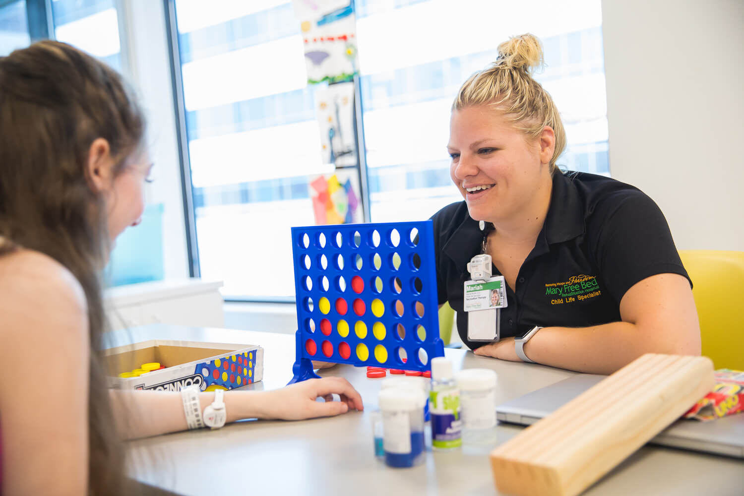 A child life specialist wearing a black short-sleeved shirt and a blond top knot plays Connect Four with a young patient. The patient's back is to the camera and the two are in a room lit with large windows. 