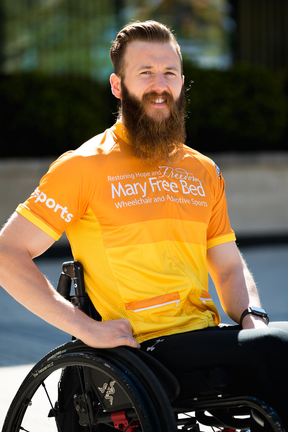 A young man is smiling. He is wearing an orange jersey that says Mary Free Bed Sports. He has red hair and a big beard, and is sitting in a black wheelchair