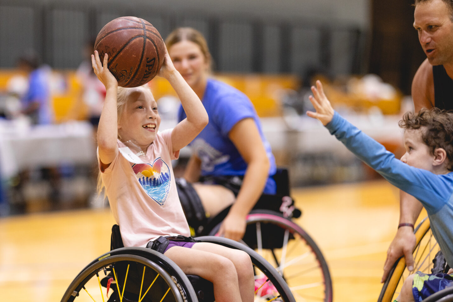 A young child in a wheelchair smiling and holding a basketball above her head during the Junior Wheelchair Sports Camp.
