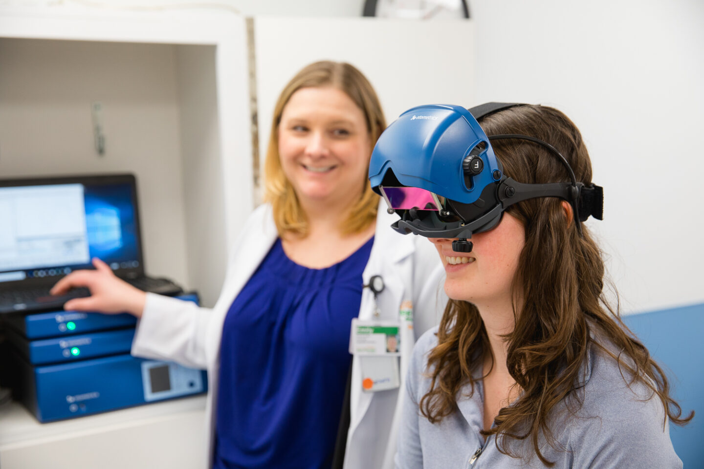 A patient wearing blue vestibular testing goggles while an audiologist operates advanced equipment.