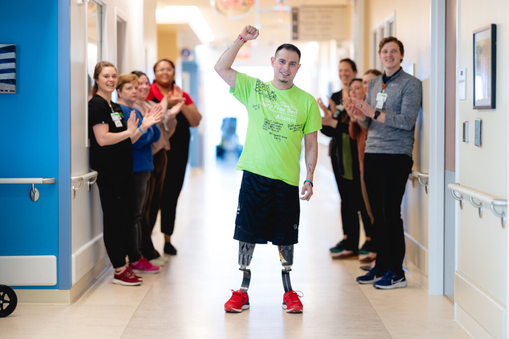 A patient with double leg prosthetics triumphantly raises his fist while walking down a hallway lined with applauding staff at Mary Free Bed.
