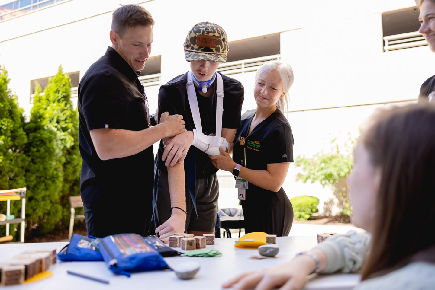 A patient using a sling support is assisted by two therapists during an outdoor recreational therapy session.