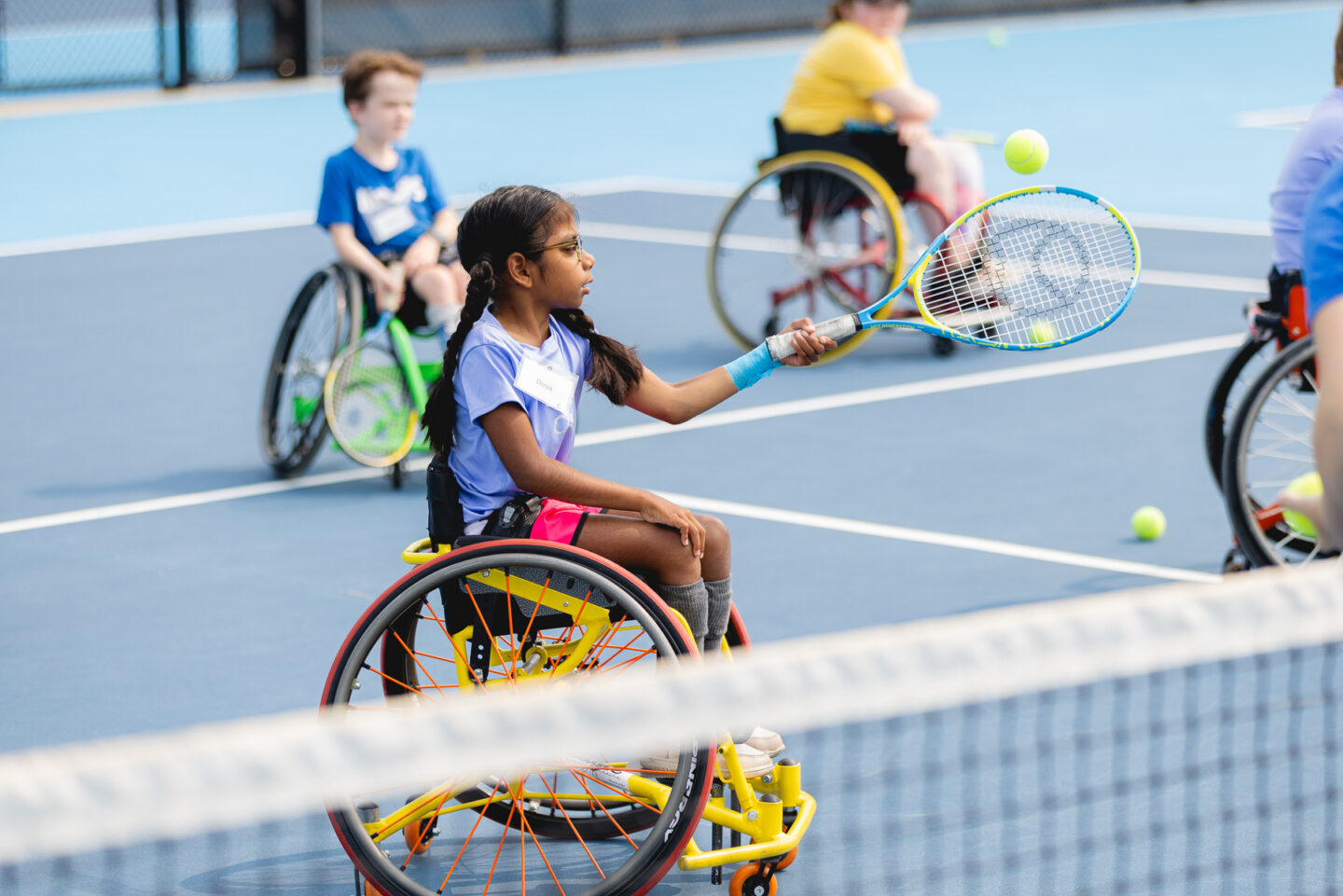 A young child in a wheelchair practicing her tennis skills on a blue court.