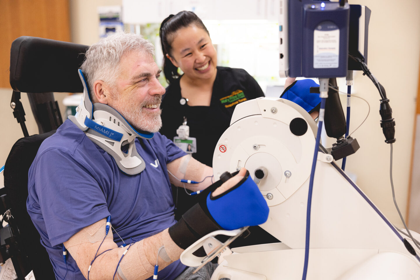 Patient with a spinal cord injury engages in therapy using a Restorative Therapies FES cycle, smiling with encouragement from the occupational therapist.