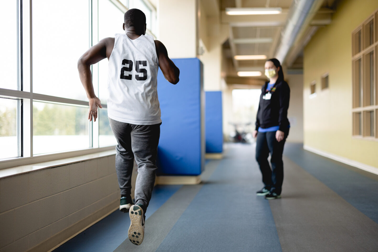 A patient performs a sprint along an indoor track at the SpartanNash YMCA while a physical therapist observes. The rehabilitation session emphasizes speed and endurance as part of sports injury recovery.