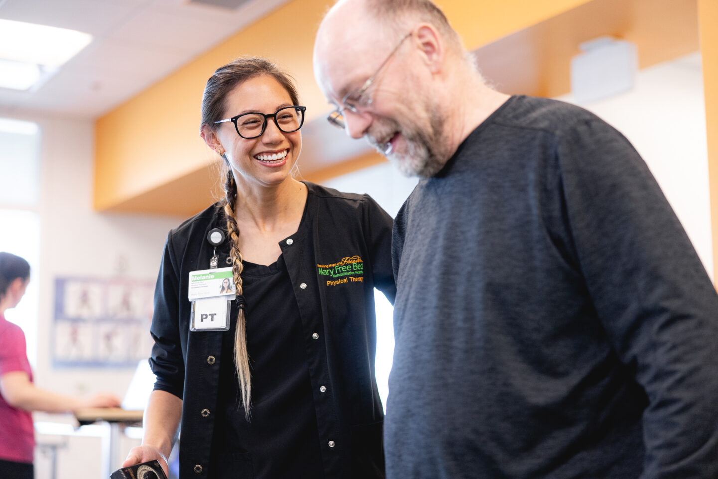 A physical therapist smiles and supports a patient during a rehabilitation session at the SpartanNash YMCA. The patient is walking with the therapist’s guidance, focusing on mobility and balance as part of his recovery process in a sports rehabilitation program.